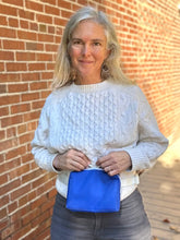 Load image into Gallery viewer, Woman wearing a white sweater holding a blue pouch against a brick wall.
