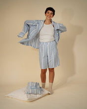Load image into Gallery viewer, A smiling man models a light blue and white striped pajama set, including a long-sleeve shirt and shorts, over a white t-shirt. A matching folded pajama set rests on a white pillow on the floor next to him, set against a solid beige studio background.
