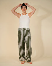 Load image into Gallery viewer, A model stands barefoot against a warm beige studio background, wearing a white sleeveless top and wide-leg trousers with vertical olive green and off-white stripes. The model has short dark hair and is holding it up with both hands.
