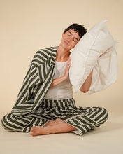 Load image into Gallery viewer, A woman sits cross-legged on a beige studio floor wearing a matching green and white striped pajama set, with a white pillow against her head.
