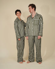 Load image into Gallery viewer, A man and a woman wearing matching striped pajama sets, standing barefoot against a plain beige studio background. The pajamas feature vertical stripes in dark green and off-white.

